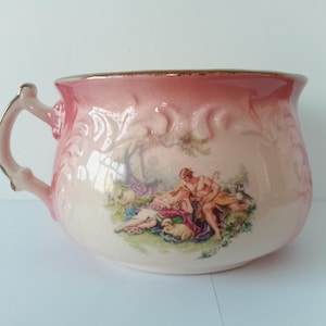 Può includere: Un vaso da notte in ceramica rosa con bordo e manico dorati. Il vaso presenta una scena dipinta di una coppia in un ambiente pastorale con pecore.