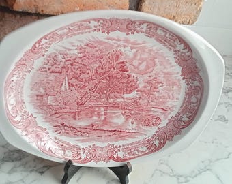 May include: A white oval serving platter with a pink and white floral design. The design features a stylized landscape with a bridge and trees.