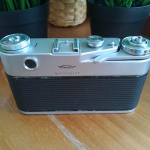 K&ouml;nnte beinhalten: Eine Vintage-Kamera in Silber und Schwarz mit einem strukturierten schwarzen Griff. Die Kamera hat silberne Drehkn&ouml;pfe und ein Objektiv. Die Seriennummer "N559023735" ist auf der Vorderseite der Kamera sichtbar. Die Kamera steht auf einer Holzoberfl&auml;che.
