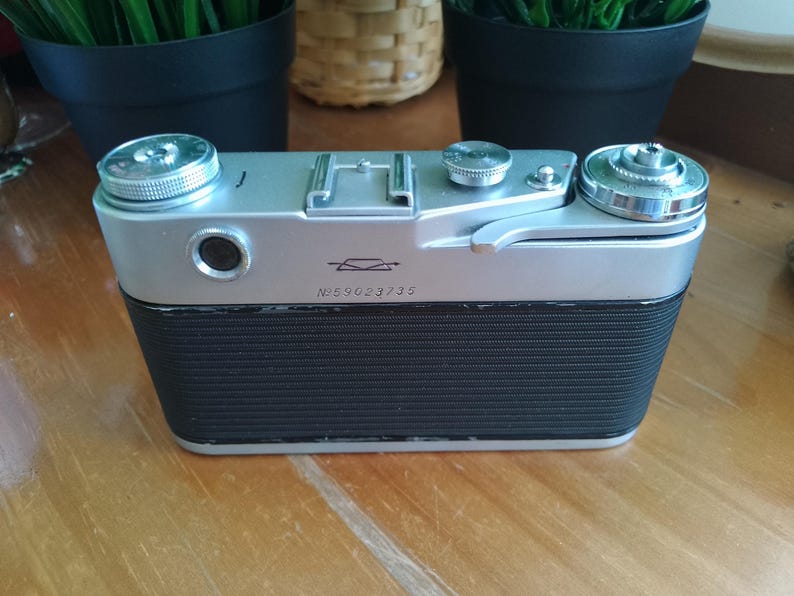 K&ouml;nnte beinhalten: Eine Vintage-Kamera in Silber und Schwarz mit einem strukturierten schwarzen Griff. Die Kamera hat silberne Drehkn&ouml;pfe und ein Objektiv. Die Seriennummer "N559023735" ist auf der Vorderseite der Kamera sichtbar. Die Kamera steht auf einer Holzoberfl&auml;che.