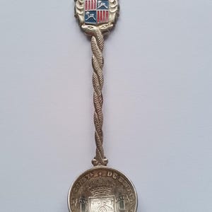 May include: Silver souvenir spoon with a twisted handle and a detailed bowl. The spoon features a crest with the word "Mallorca" and a coat of arms. The bowl is engraved with text and a crest. A decorative collectible.