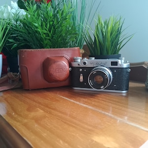 Puede incluir: Una cámara telemétrica vintage FED-2 con cuerpo plateado y negro, que presenta un objetivo circular y texto en cirílico en la placa superior. La cámara viene acompañada de un estuche protector de cuero marrón con una correa a juego.