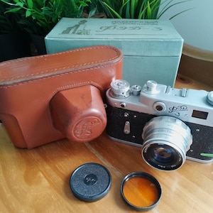 May include: A silver and black FED-3 rangefinder camera with a lens, a brown leather carrying case, a light green cardboard box with Cyrillic text, a black lens cap, and a circular orange filter. The camera body features the text FED 3.
