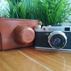 Puede incluir: Una cámara vintage FED-2 con cuerpo negro y detalles plateados, junto a una funda de cuero marrón. La lente de la cámara es visible y la funda tiene una tapa de lente a juego. Un artículo de fotografía clásico.