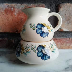Può includere: Un set di lattiera e zuccheriera in ceramica bianca con un motivo floreale blu e giallo. La lattiera ha un manico e un beccuccio. La zuccheriera ha un coperchio.