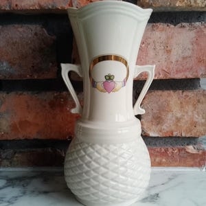 Puede incluir: Jarrón de cerámica color crema con diseño Claddagh. El jarrón presenta una parte superior ancha y acanalada, dos asas curvas y una base texturizada con patrón de diamantes. El emblema de Claddagh incluye un corazón, una corona y manos, que simbolizan el amor, la lealtad y la amistad.