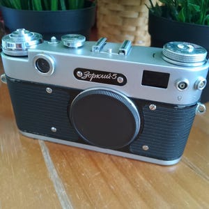 K&ouml;nnte beinhalten: Eine Vintage-Kamera in Silber und Schwarz mit dem Text "Зоркий 5" auf der Vorderseite. Die Kamera hat einen schwarzen Objektivdeckel und verschiedene Kn&ouml;pfe und Schalter. Die Kamera steht auf einer Holzoberfl&auml;che.