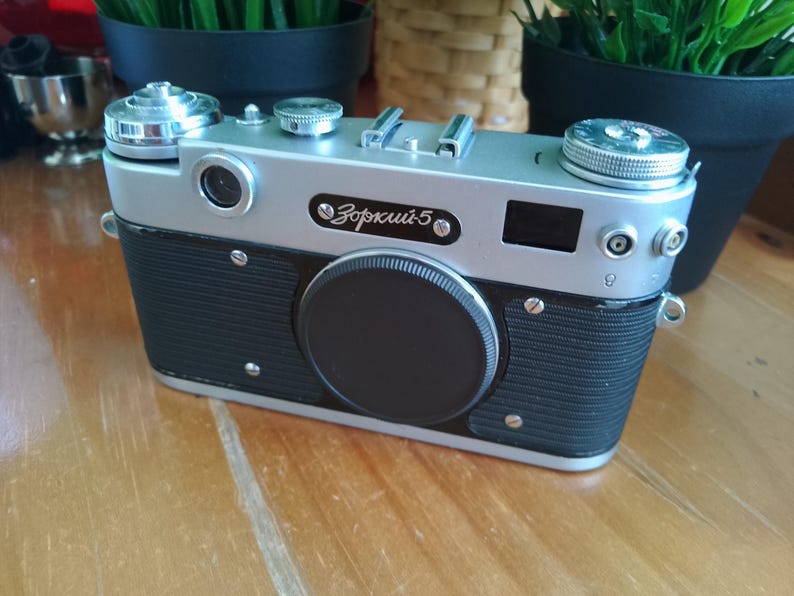 K&ouml;nnte beinhalten: Eine Vintage-Kamera in Silber und Schwarz mit dem Text "Зоркий 5" auf der Vorderseite. Die Kamera hat einen schwarzen Objektivdeckel und verschiedene Kn&ouml;pfe und Schalter. Die Kamera steht auf einer Holzoberfl&auml;che.