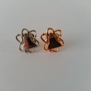 Könnte beinhalten: Zwei glockenförmige Anstecknadeln. Eine ist silberfarben, die andere goldfarben. Jede Anstecknadel hat eine Glockenform mit dekorativer Kontur. Die Glocken sind in einem stilisierten Blumendesign zentriert.