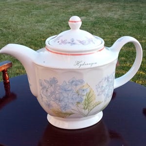 Può includere: Teiera in ceramica bianca con coperchio, manico e beccuccio. La teiera è decorata con fiori di ortensia blu e foglie verdi. La parola "Hydrangea" è scritta sul lato. La teiera è appoggiata su un tavolo marrone scuro.