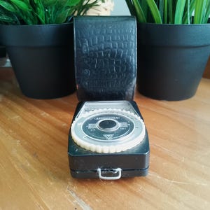 May include: A vintage black light meter featuring a textured, crocodile-patterned flip-up cover and a circular dial with white markings. The device has a central black button, a silver metal loop at the base, and Cyrillic text on the face.