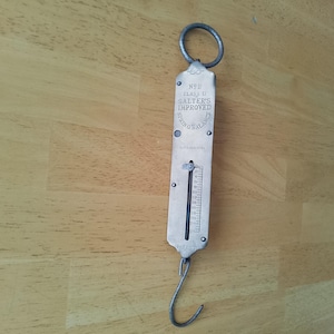 Op de afbeelding: Vintage Salter's Improved veerschaal. De metalen weegschaal heeft een haak aan de onderkant en een ring aan de bovenkant. Het heeft een meetindicator en de tekst "Salter's Improved Spring Balance".