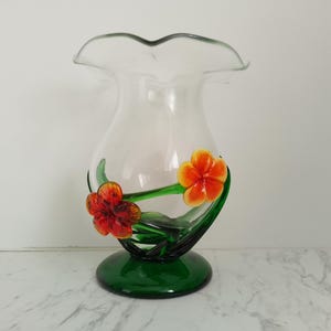 May include: A clear glass vase with a wide mouth and a green base. The vase is decorated with three dimensional glass flowers in shades of orange, red, and green.
