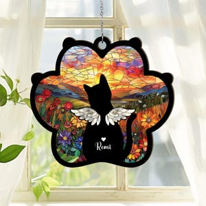 Könnte beinhalten: Schwarze Katzensilhouette mit Engelsflügeln, vor einer Sonnenuntergangsszene im Glasmalerei-Stil. Die Katze befindet sich in einem pfotenförmigen Rahmen mit dem Namen "Remi" und einem Herzen. Dekoratives Hängeornament.