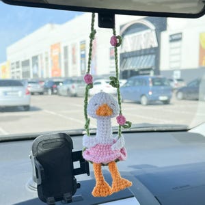 Op de afbeelding: Een witte gehaakte gans die een roze jurk draagt en op een groene schommel zit met roze bloemen. De gans hangt aan de achteruitkijkspiegel van een auto.