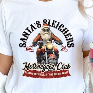 Puede incluir: Camiseta blanca con un gr&aacute;fico de Pap&aacute; Noel conduciendo una motocicleta. El gr&aacute;fico dice "Santa's Sleighers est. 24 Motorcycle Club Decking the halls, hitting the highways".