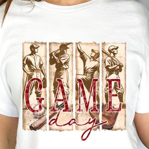 K&ouml;nnte beinhalten: Wei&szlig;es T-Shirt mit Baseball-Motiv. Das Design zeigt vier Illustrationen von Baseballspielern im Vintage-Stil und die Worte "GAME DAY" in einer verblassten, roten Schriftart. Das T-Shirt wird von einer Person getragen.