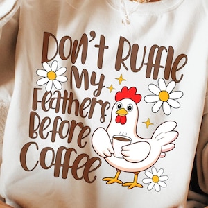 May include: Cream-colored sweatshirt with the text "Don't Ruffle My Feathers Before Coffee" in brown. A cartoon chicken holding a coffee cup is featured, along with daisy and star graphics. The sweatshirt is a casual, comfortable style.