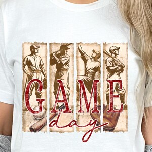 K&ouml;nnte beinhalten: Wei&szlig;es T-Shirt mit einem Baseball-Motiv. Das Design zeigt vier Baseballspieler und die Worte "GAME DAY" in einer roten, strukturierten Schriftart. Das T-Shirt hat einen klassischen Rundhalsausschnitt.