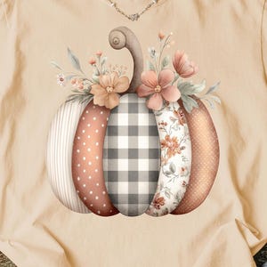Puede incluir: Una camiseta beige Comfort Colors con un diseño de calabaza en acuarela. La calabaza está segmentada con varios patrones, incluyendo lunares, cuadros vichy e estampados florales, y está adornada con flores y hojas. La camiseta tiene cuello redondo.