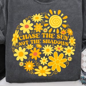 May include: Dark gray sweatshirt with a floral design in yellow and orange. The design includes a sun and the text "CHASE THE SUN NOT THE SHADOWS". The sweatshirt has long sleeves and a crew neck.