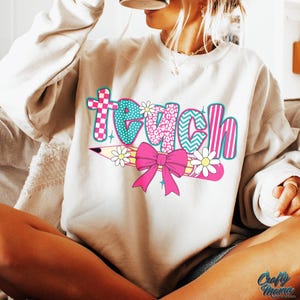 May include: A white sweatshirt with the word "teach" printed on it in colorful letters. The letters are decorated with different patterns, including polka dots, stripes, and animal print. A pink bow and a pencil with a flower are also included in the design.