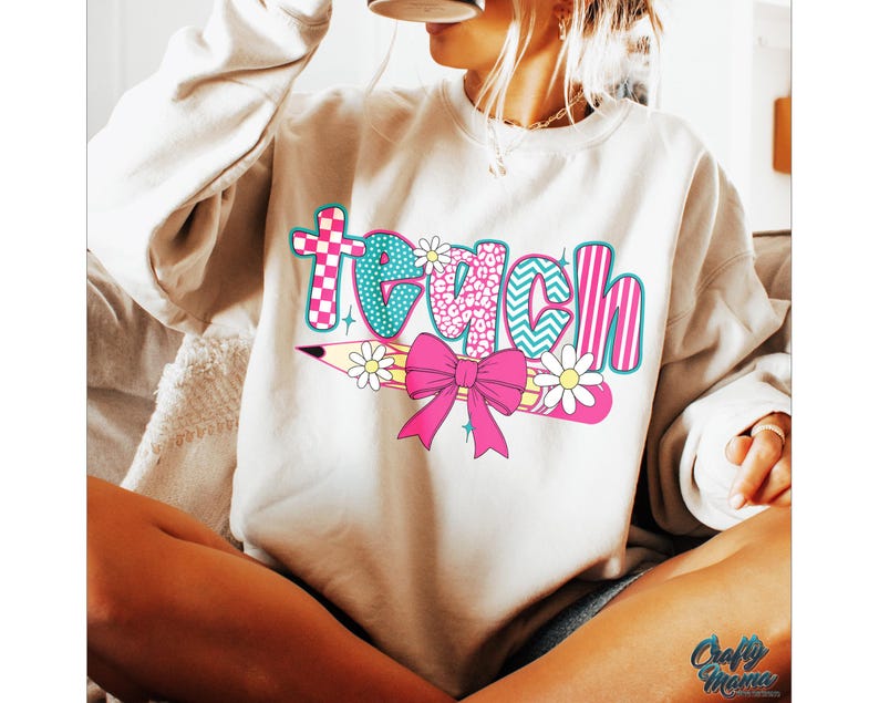 May include: A white sweatshirt with the word "teach" printed on it in colorful letters. The letters are decorated with different patterns, including polka dots, stripes, and animal print. A pink bow and a pencil with a flower are also included in the design.