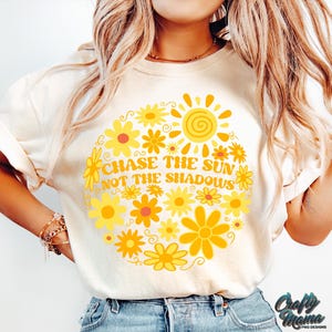May include: Cream-colored t-shirt featuring a circular floral design with a sun graphic. The design includes yellow flowers and the text "CHASE THE SUN NOT THE SHADOWS". The shirt has short sleeves and a crew neck.