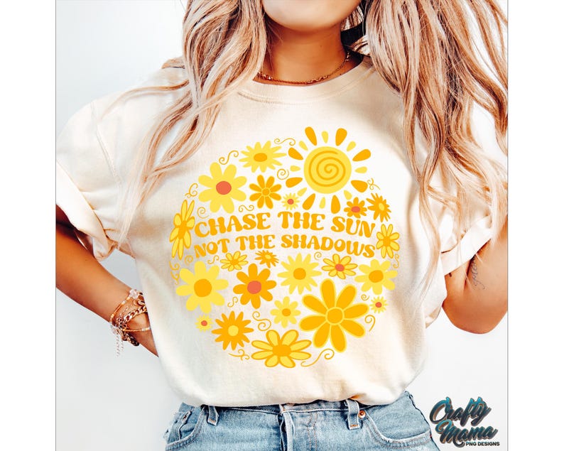 May include: Cream-colored t-shirt featuring a circular floral design with a sun graphic. The design includes yellow flowers and the text "CHASE THE SUN NOT THE SHADOWS". The shirt has short sleeves and a crew neck.