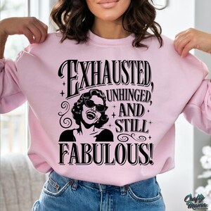 May include: A pale pink sweatshirt featuring the words "EXHAUSTED, UNHINGED, AND STILL FABULOUS!" in black. The design includes a black and white illustration of a woman in sunglasses. The sweatshirt has a crew neck.