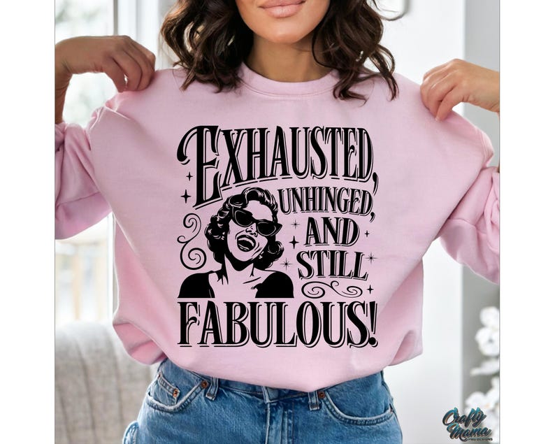 May include: A pale pink sweatshirt featuring the words "EXHAUSTED, UNHINGED, AND STILL FABULOUS!" in black. The design includes a black and white illustration of a woman in sunglasses. The sweatshirt has a crew neck.