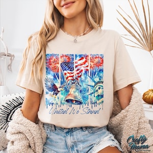Pu&ograve; includere: Una maglietta beige con un design ad acquerello che presenta una bandiera americana, una campana, un'aquila calva e la Statua della Libert&agrave;. Il design include anche fuochi d'artificio e il testo "United We Stand".
