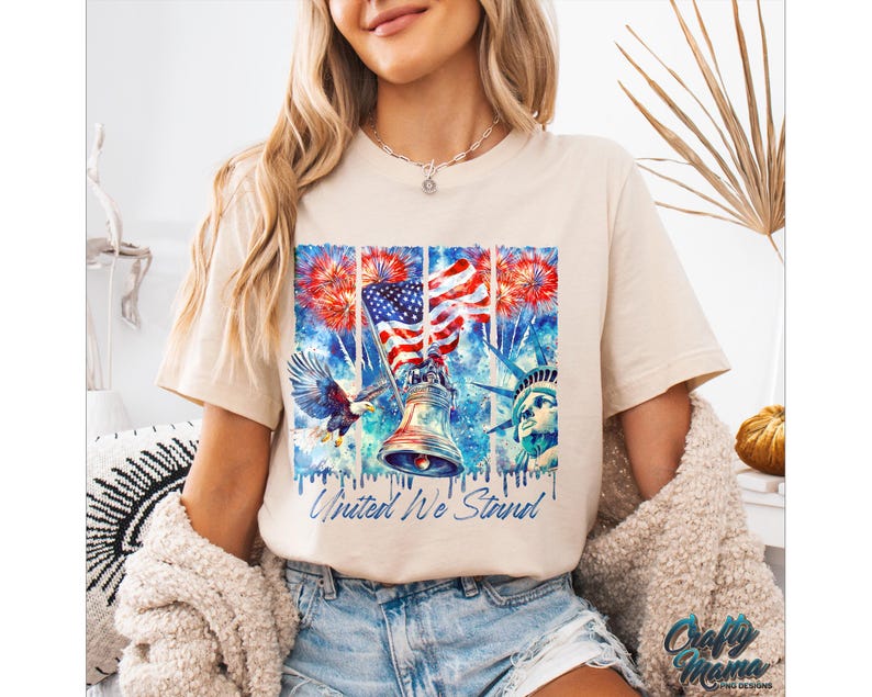 Pu&ograve; includere: Una maglietta beige con un design ad acquerello che presenta una bandiera americana, una campana, un'aquila calva e la Statua della Libert&agrave;. Il design include anche fuochi d'artificio e il testo "United We Stand".