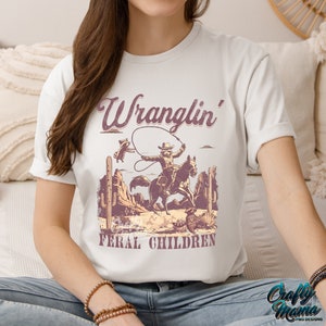 May include: Off-white t-shirt featuring a vintage-style graphic of a cowboy riding a horse, roping a raccoon, with the text "Wranglin' Feral Children." The design includes cacti and desert scenery. The shirt is a casual wear item.