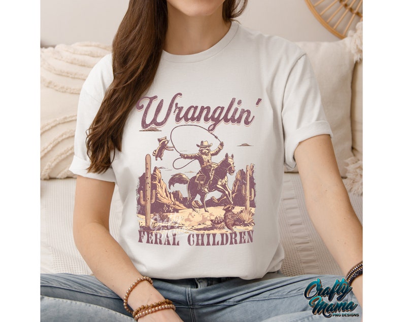 May include: Off-white t-shirt featuring a vintage-style graphic of a cowboy riding a horse, roping a raccoon, with the text "Wranglin' Feral Children." The design includes cacti and desert scenery. The shirt is a casual wear item.