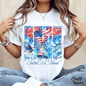 Pu&ograve; includere: Maglietta bianca con un design ad acquerello di una bandiera americana, fuochi d'artificio, la Statua della Libert&agrave; e la Liberty Bell. Il testo "United We Stand" &egrave; stampato sulla maglietta.