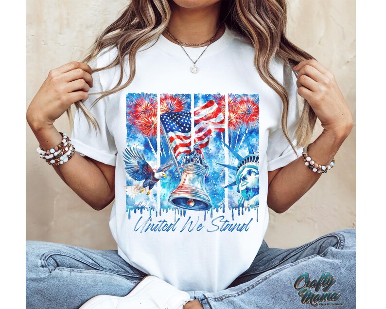 Pu&ograve; includere: Maglietta bianca con un design ad acquerello di una bandiera americana, fuochi d'artificio, la Statua della Libert&agrave; e la Liberty Bell. Il testo "United We Stand" &egrave; stampato sulla maglietta.