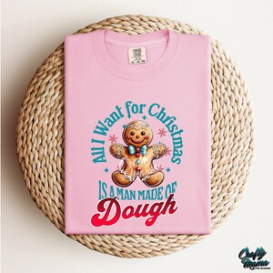 May include: A light pink t-shirt with the text "All I want for Christmas is a man made of dough" in a circle around a gingerbread man graphic. The gingerbread man has a blue bow tie and colorful candy buttons.
