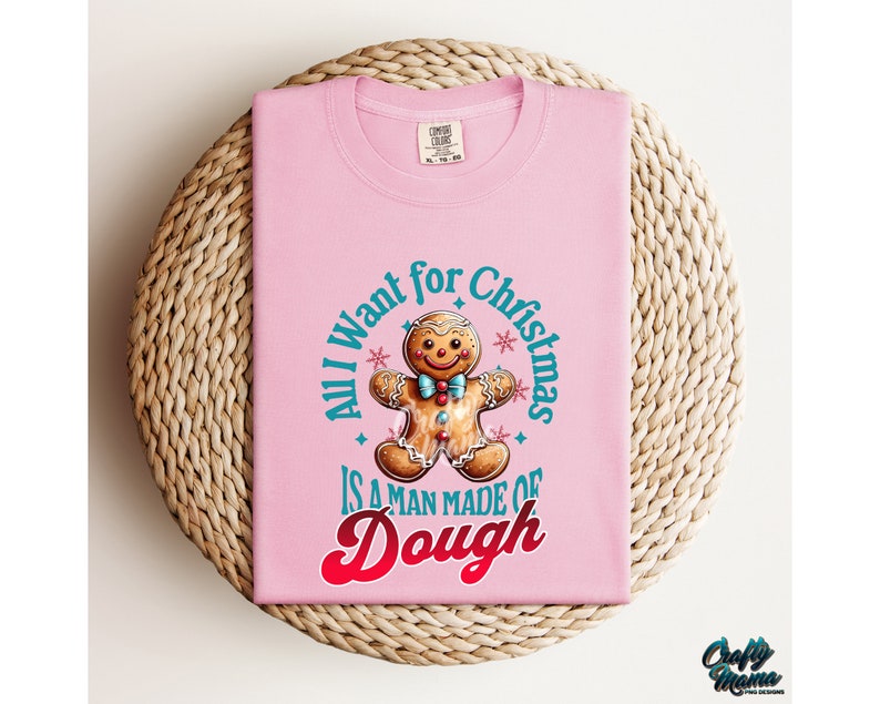 May include: A light pink t-shirt with the text "All I want for Christmas is a man made of dough" in a circle around a gingerbread man graphic. The gingerbread man has a blue bow tie and colorful candy buttons.