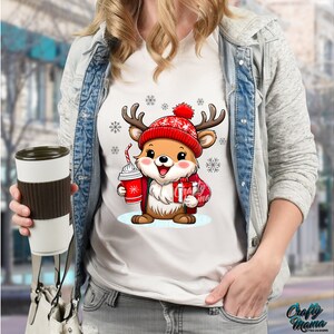 May include: White t-shirt with a cartoon reindeer wearing a red and white Santa hat and holding a cup of hot chocolate and a gift. The reindeer is surrounded by snowflakes.