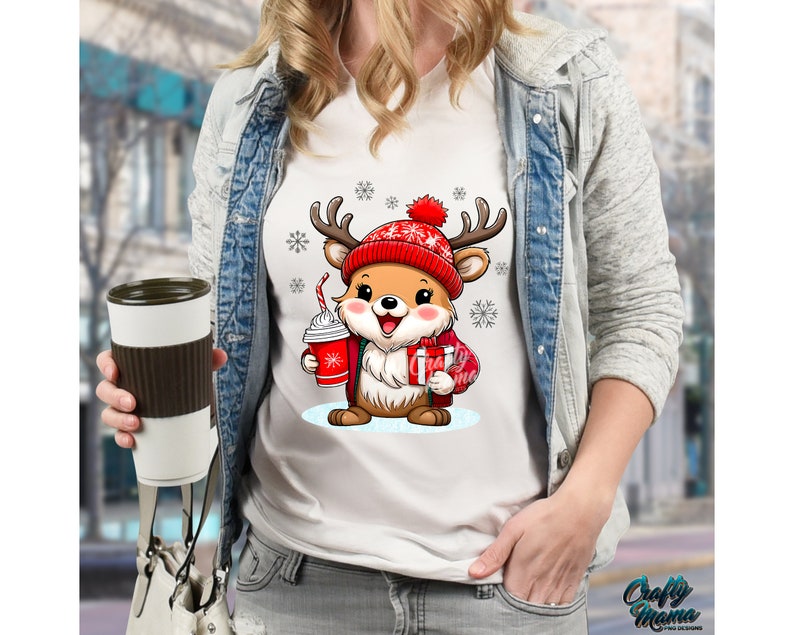 May include: White t-shirt with a cartoon reindeer wearing a red and white Santa hat and holding a cup of hot chocolate and a gift. The reindeer is surrounded by snowflakes.