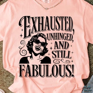 May include: A pale pink t-shirt featuring the text "EXHAUSTED, UNHINGED, AND STILL FABULOUS!" in black. The design includes a black and white illustration of a woman wearing sunglasses. The t-shirt has short sleeves and a round neckline.