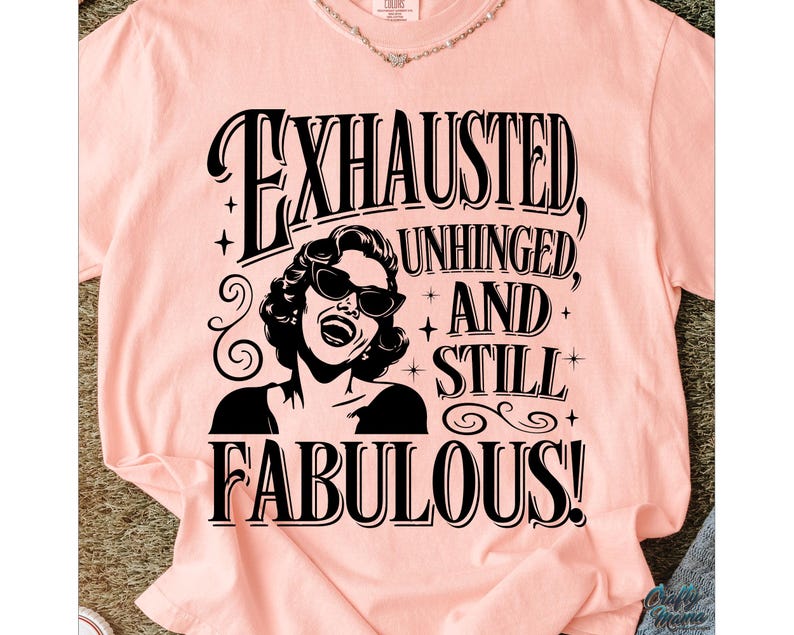 May include: A pale pink t-shirt featuring the text "EXHAUSTED, UNHINGED, AND STILL FABULOUS!" in black. The design includes a black and white illustration of a woman wearing sunglasses. The t-shirt has short sleeves and a round neckline.