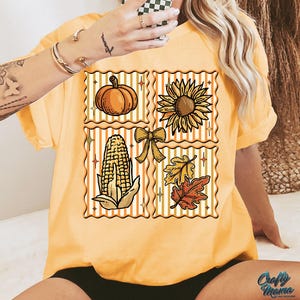 Puede incluir: Camiseta amarilla mostaza con un dise&ntilde;o de tem&aacute;tica oto&ntilde;al. El dise&ntilde;o presenta una calabaza, un girasol, ma&iacute;z, un lazo y hojas dispuestas en un patr&oacute;n de cuadr&iacute;cula. La camiseta tiene mangas cortas y un corte holgado. El texto "Crafty Mama" es visible en la parte inferior derecha.