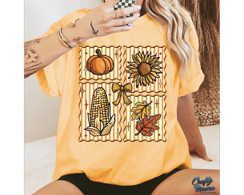 Puede incluir: Camiseta amarilla mostaza con un dise&ntilde;o de tem&aacute;tica oto&ntilde;al. El dise&ntilde;o presenta una calabaza, un girasol, ma&iacute;z, un lazo y hojas dispuestas en un patr&oacute;n de cuadr&iacute;cula. La camiseta tiene mangas cortas y un corte holgado. El texto "Crafty Mama" es visible en la parte inferior derecha.
