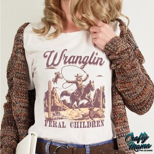 May include: Cream-colored t-shirt with a vintage-style graphic of a cowboy roping a raccoon, with the words "Wranglin' Feral Children." The design features a desert landscape in shades of brown and purple.