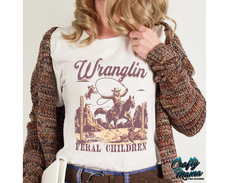 May include: Cream-colored t-shirt with a vintage-style graphic of a cowboy roping a raccoon, with the words "Wranglin' Feral Children." The design features a desert landscape in shades of brown and purple.