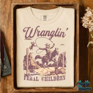 May include: Cream-colored t-shirt with a graphic featuring a cowboy riding a horse, roping a raccoon, and the text "Wranglin' Feral Children." The design includes desert scenery in shades of purple and brown.