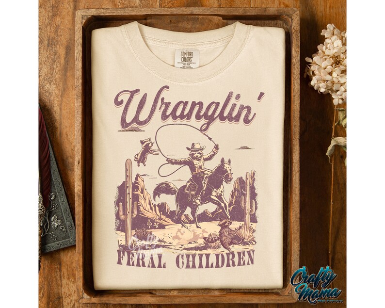 May include: Cream-colored t-shirt with a graphic featuring a cowboy riding a horse, roping a raccoon, and the text "Wranglin' Feral Children." The design includes desert scenery in shades of purple and brown.