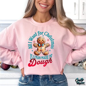 May include: A light pink sweatshirt with the text "All I want for Christmas is a man made of dough" and a gingerbread man graphic. The graphic is in shades of brown, blue, and red. The sweatshirt is a crew neck style.